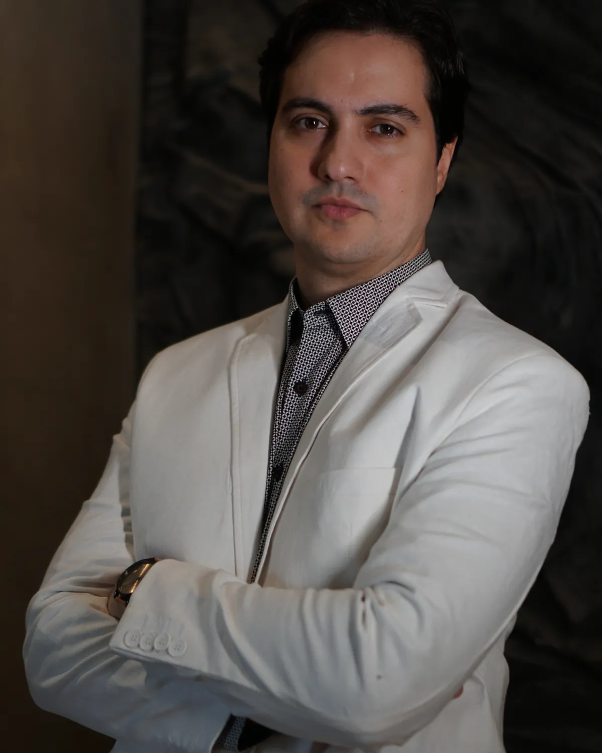 Dr. Diego Paiva de blazer claro e braços cruzados, em estúdio com luz quente
